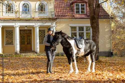 Icelandic horse and female rider in autumn scenery with maple leaf on the ground. Rider wearing black helmet. Old house in the background.