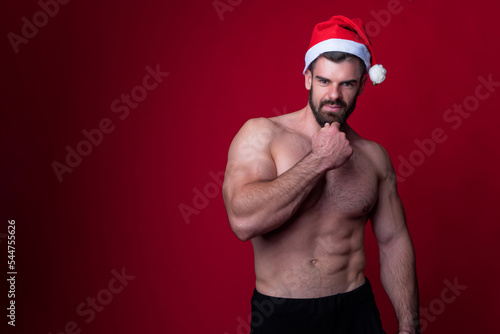 Sexy santa, young shirtless bodybuilder wearing a christmas hat, playing santa claus for holidays, red background studio portrait © Teodor Lazarev