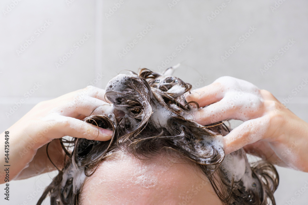 A girl washes her hair with shampoo on gray tiles background, front view. - obrazy, fototapety, plakaty 