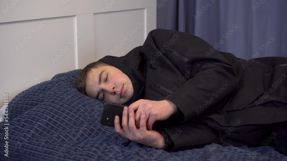 A young man in a jacket lies on the bed and uses a smartphone. The man did not pay the bills and was left without heating in the winter. A young man in a jacket lies on the bed and uses a smartphone. The man did not pay the bills and was left without heating in the winter.