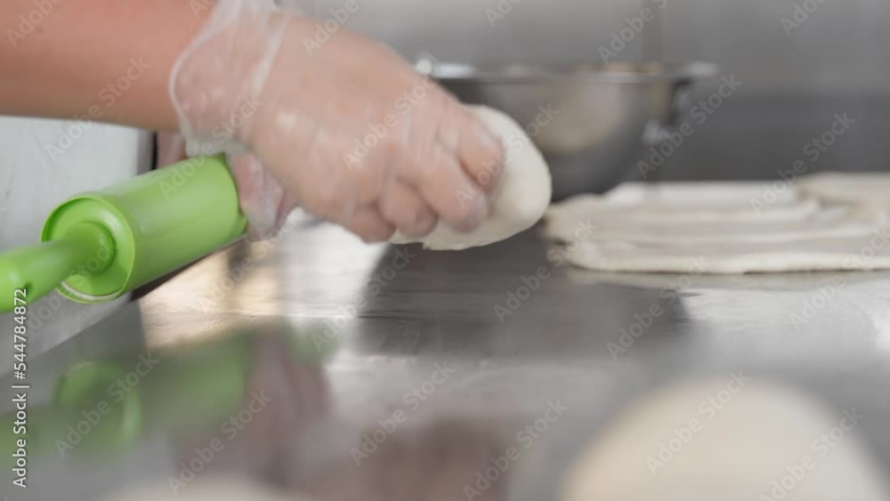 The chef uses a rolling pin to stretch raw dough in the kitchen. Rolling pin expanding raw dough in a bakery. A gloved worker finishes lengthening the dough with his hands. The chef uses a rolling pin to stretch raw dough in the kitchen. Rolling pin expanding raw dough in a bakery. A gloved worker finishes lengthening the dough with his hands.