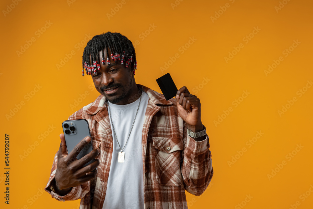 Obraz premium Smiling young african man on yellow background using mobile phone and holding credit card.
