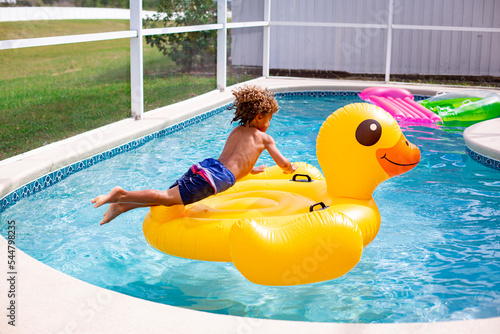Young diverse little boy jumping onto a large inflatable pool toy in a backyard swimming pool on a warm summer day. Action photo of the child jumping through the air