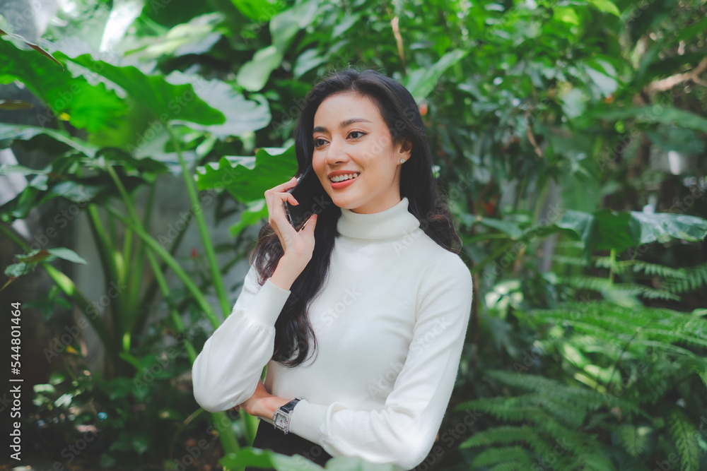 A young smart business woman, freelance or architect is happy while talking with customer by mobile phone in outdoor natural coffee shop. Concept of communication and conversation, relaxing working. A young smart business woman, freelance or architect is happy while talking with customer by mobile phone in outdoor natural coffee shop. Concept of communication and conversation, relaxing working.