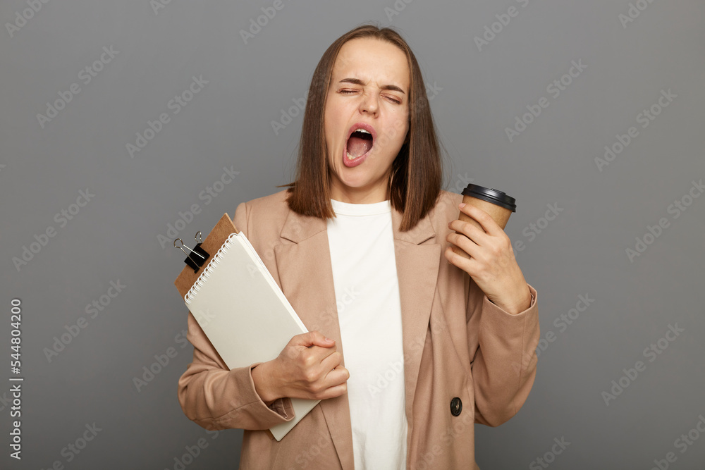 Portrait of tired overworked brown-haired woman wearing jacket holding clipboard with documents and hot coffee to go, needs to wake up, isolated over gray background. Portrait of tired overworked brown-haired woman wearing jacket holding clipboard with documents and hot coffee to go, needs to wake up, isolated over gray background.