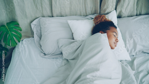Fotografia Young African American woman is sleeping in comfortable bed under warm blanket having rest on beautiful linen