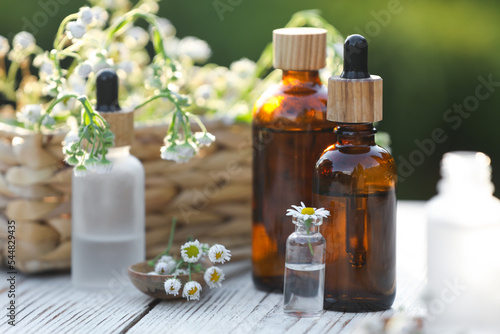 Bottles of chamomile essential oil and flowers on white wooden table, closeup