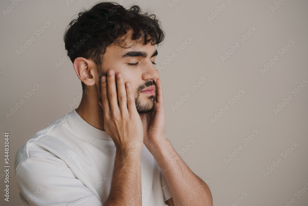 Side view of a confused young man touching his face standing against grey background with copy space. Skin care. Body care. Side view of a confused young man touching his face standing against grey background with copy space. Skin care. Body care.
