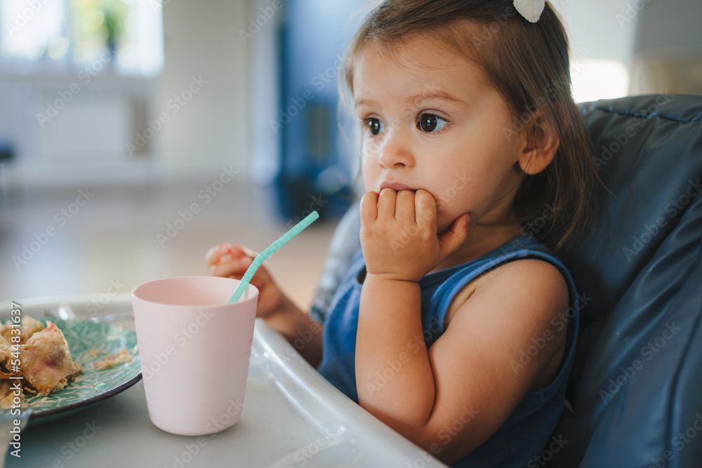 Pensive sweet baby girl sitting in highchair with food. First solid food or child care concept. Healthy eating. Happy family. Pensive sweet baby girl sitting in highchair with food. First solid food or child care concept. Healthy eating. Happy family.