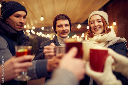 Happy smiling friends with cups of mulled wine having fun  spending time together at winter fair at evening time. Winter holidays  Christmas concept
