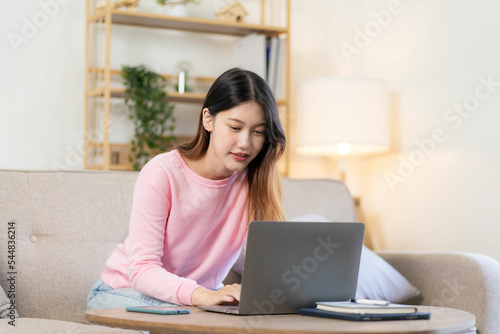 Asian woman smiling and working from home on laptop online to internet on living room sofa. At home with the concept and lifestyle
