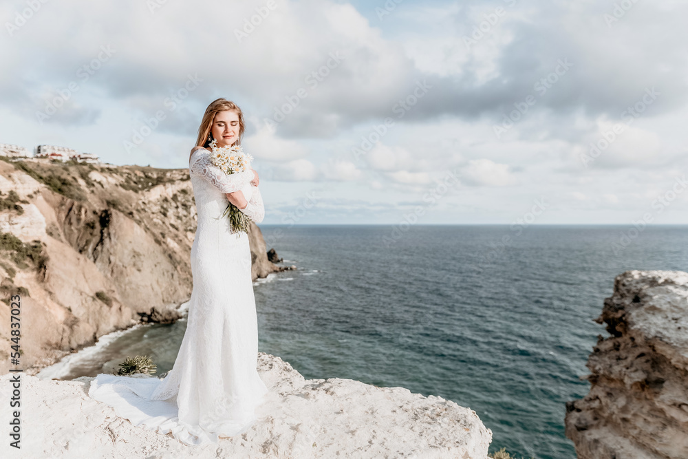 Romantic bride, blond girl in white wedding dress with open shoulders posing with sea and rocks in background. Stylish young woman standing on cliff's edge and looking into a ocean mountains at sunset Romantic bride, blond girl in white wedding dress with open shoulders posing with sea and rocks in background. Stylish young woman standing on cliff's edge and looking into a ocean mountains at sunset