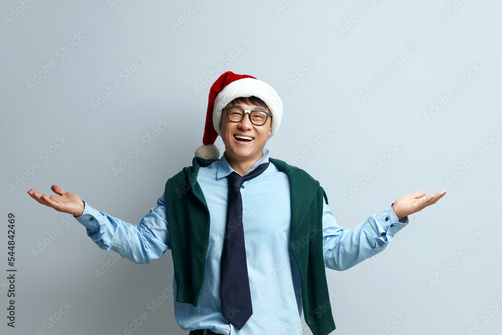 Shrugging Man Smiling Studio. Confused Guy in Santa Claus Hat Standing with No Idea Gesture, Shrugging Shoulders Raising Hands. Indoor Studio Shot Isolated on Grey Background 
