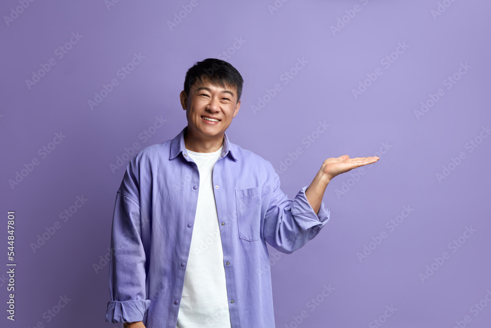 Smiling Man Pointing Hand. Positive Asian Guy Pointing Away Paying Your Attention at Empty Space for Advertisement. Indoor Studio Shot Isolated on Violet Background Smiling Man Pointing Hand. Positive Asian Guy Pointing Away Paying Your Attention at Empty Space for Advertisement. Indoor Studio Shot Isolated on Violet Background