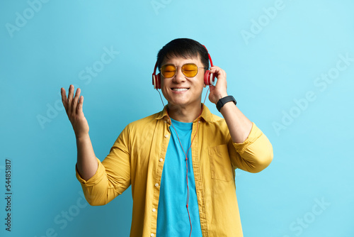 Asian Man Listening Music. Positive Man Listening Favourite Song Wearing Headphones. Indoor Studio Shot Isolated on Blue Background 