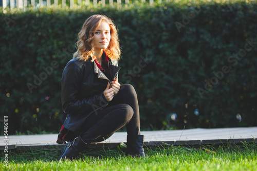 Beautiful young woman in a black leather jacket sits on a boardwalk. Outdoor portrait in city park. Copy space