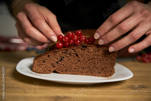 Woman with a passion for cooking decorates a slice of handmade chocolate cake in her home kitchen with blackberries and red currants.