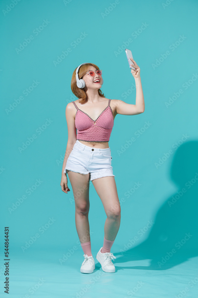 Full length image of pleased woman dancing and listening music by the smartphone over cyan background Full length image of pleased woman dancing and listening music by the smartphone over cyan background