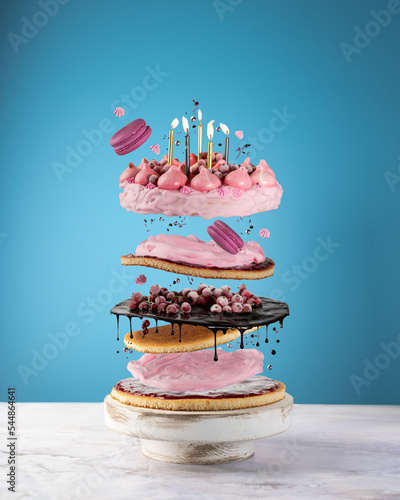 Flying cake layers with berries,chocolate,macarons,pink cream,festive burning candles on blue background.Birthday cake in freeze motion,food levitation.