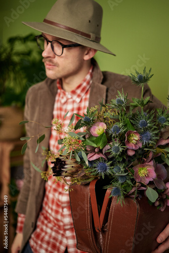 Attractive vintage styled male person in eyeglasses, velvet jacket and brown hat staying with flower bouquet and briefcase in flower store. High quality vertical image © boytsov