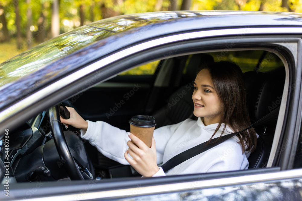 Portrait of a young and cheerful woman with coffee cup driving luxury car in the city. Smiling pretty young woman drinking take out coffee when driving to work in the morning