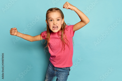 Portrait of little beautiful girl, child in pink T-shirt posing, aggressively running isolated over blue studio background