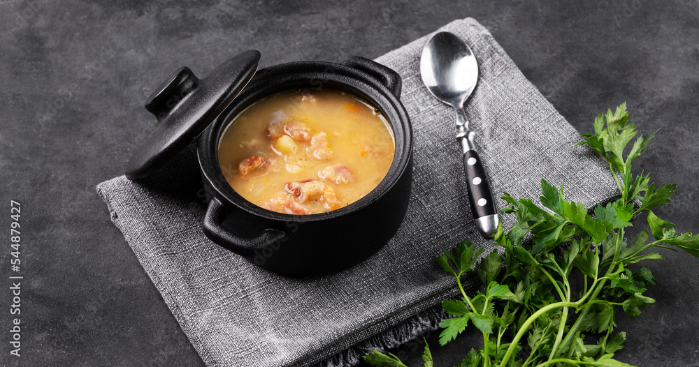 Pea soup with smoked ham in black soucepan on dark background with herb. Pea soup with smoked ham in black soucepan on dark background with herb.