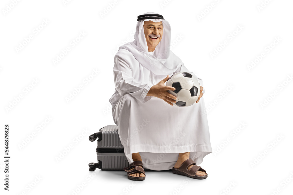 Fototapeta premium Smiling mature arab man in ethnic clothes sitting on a suitcase and holding a football