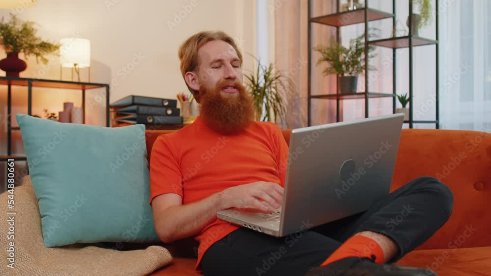 Handsome redhead man freelancer at home living room sitting on orange couch, opens laptop start working. Young guy works on notebook, sends messages, makes online purchases, watching movies, working Handsome redhead man freelancer at home living room sitting on orange couch, opens laptop start working. Young guy works on notebook, sends messages, makes online purchases, watching movies, working