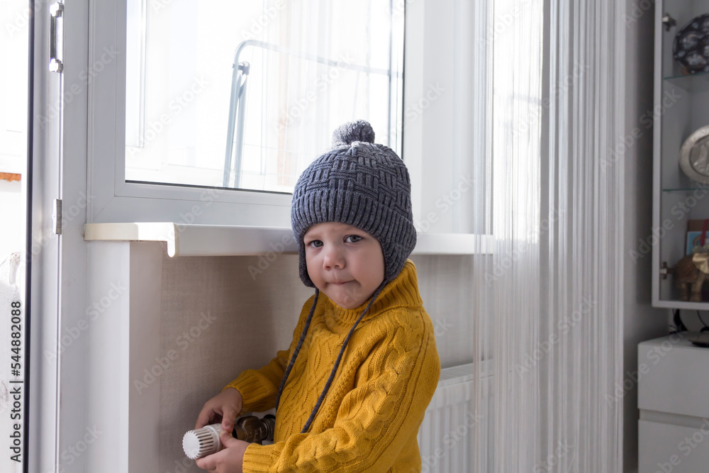 A small child in a yellow sweater and a gray hat is standing near a heater with a thermostat.,, A small child in a yellow sweater and a gray hat is standing near a heater with a thermostat.,,