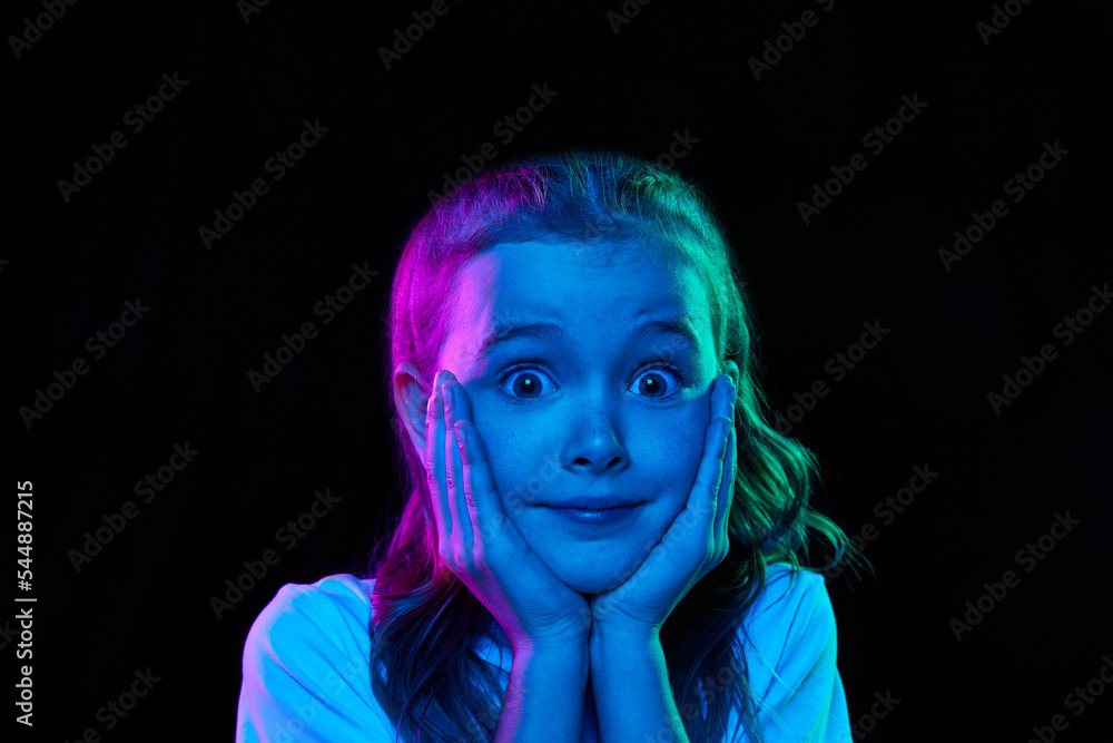 Portrait of little beautiful girl, child in white T-shirt posing isolated over black studio background in neon light. Cute kid