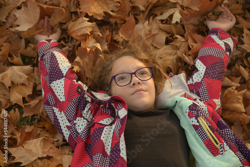 A younger girl with glasses on her head, lying in the park on the autumn yellow leaves and looking at the sky with outstretched arms.