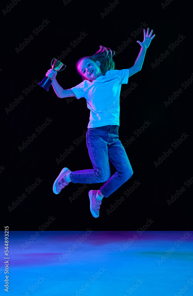 Portrait of little beautiful girl, child in white T-shirt with trophy and jumping isolated over black studio background in neon light
