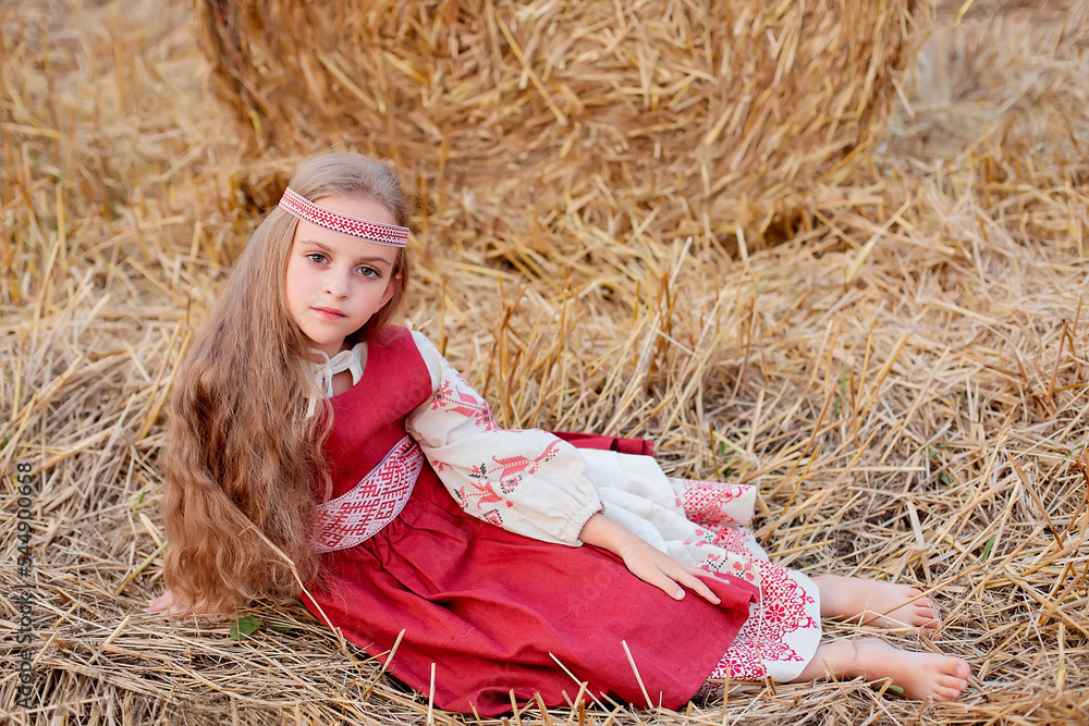a blonde girl with big brown eyes in a Slavic costume is sitting on a field by a haystack a blonde girl with big brown eyes in a Slavic costume is sitting on a field by a haystack