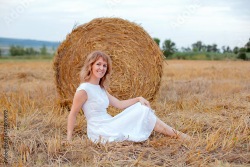 a young happy girl in a white dress is sitting by a haystack field nature