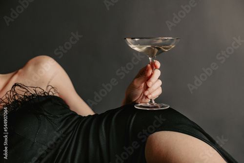 Unrecognizable woman in evening black dress, lying on a black studio background and holding glass of champagne in her hands