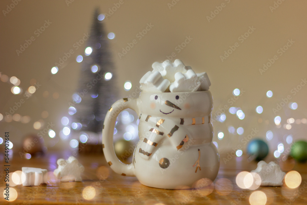 a snowman cup filled with white marshmallow snowflakes on a table against a background of a Christmas tree and holiday lights