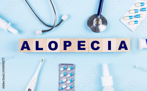 On a blue background, a stethoscope, a thermometer, other medicines and wooden cubes with the text ALOPECIA. View from above. Medical concept