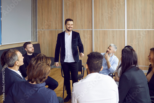 Cheerful businessman standing talking to colleagues who are sitting in circle around him in office. People sitting in circle on chairs share ideas and discuss project during brainstorming session.