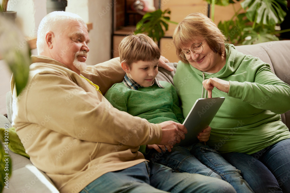 Life portrait of friendly family, grandparent and their grandson sitting on sofa and spending time together, using modern gadgets, talking, studying