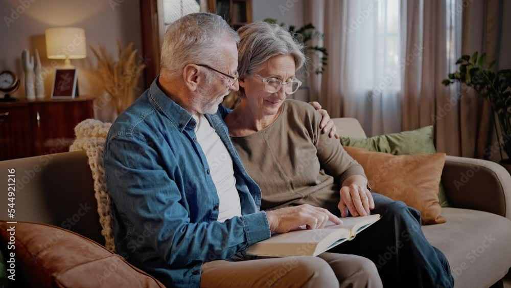 Happy elderly couple reads book together sitting on sofa Happy elderly couple reads book together sitting on sofa