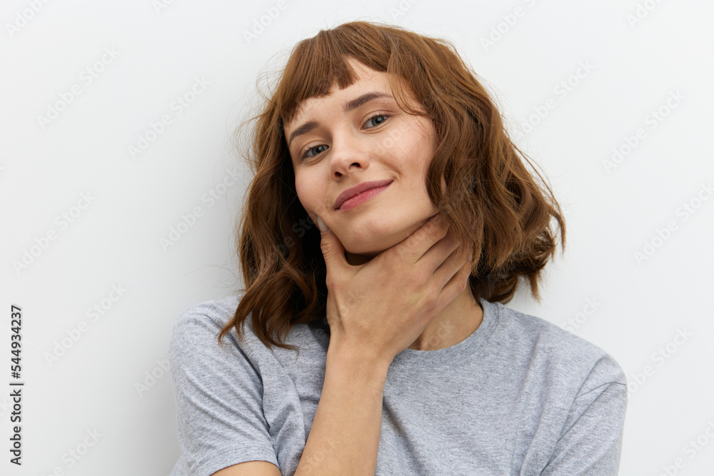 a beautiful, funny, attractive woman in a grey cotton T-shirt stands on a light background and smiles broadly with her hand on her neck a beautiful, funny, attractive woman in a grey cotton T-shirt stands on a light background and smiles broadly with her hand on her neck