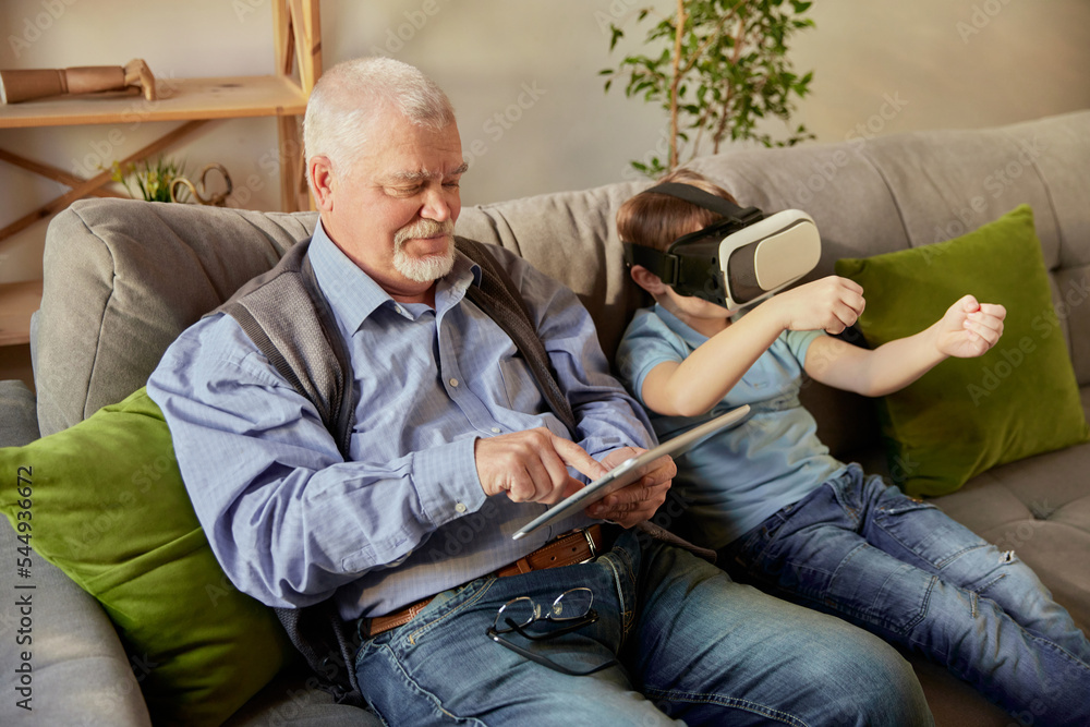 Old man and little boy, grandfather and grandson in casual clothes sitting on sofa at home and using VR glasses. Concept of hobbies, leisure activities, family