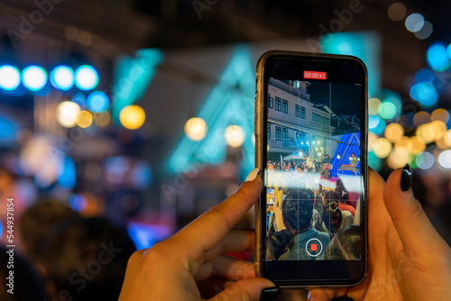 Konstfotografi Hands of unrecognizable person holding his mobile with both hands while videotaping a nightly street music show