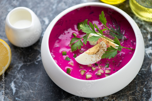 Cold beetroot soup with slices of boiled chicken egg and fresh greens served in a white bowl, horizontal shot, middle closeup