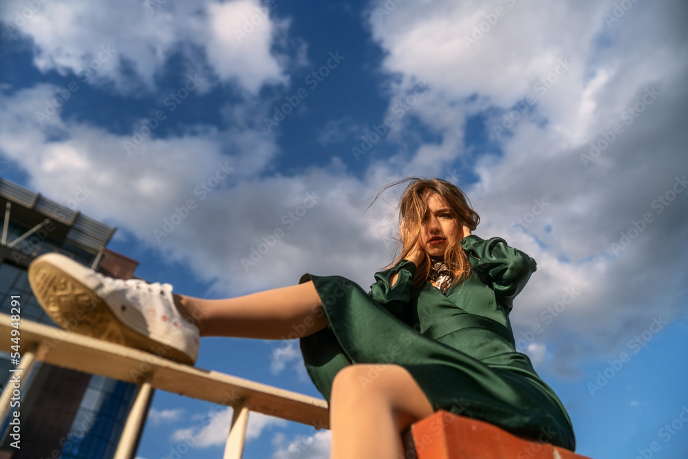 Street style fashion portrait of a cute young brown haired girl in a green dress under the cloudy sky. Attractive slim woman sits on the fence and poses in the light of the sun.