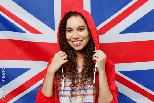Young smiling happy cheerful fun foreign woman of African American ethnicity 20s wear red raincoat jacket look camera isolated on Great Britain flag background Wet fall British weather season concept