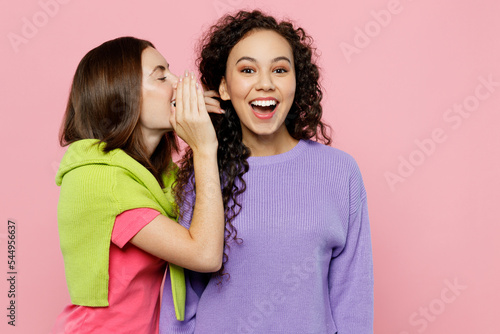 Young two friends fun women 20s wears green purple shirts together whispering gossip and tells secret behind her hand sharing news isolated on pastel plain light pink color background studio portrait