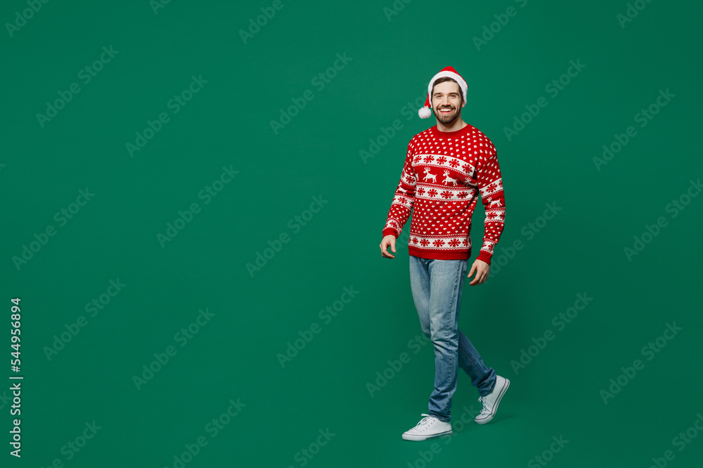 Full body merry smiling young man 20s wear red warm knitted sweater Santa hat posing walking going look camera isolated on plain dark green background studio New Year 2023 holiday celebration concept Full body merry smiling young man 20s wear red warm knitted sweater Santa hat posing walking going look camera isolated on plain dark green background studio New Year 2023 holiday celebration concept