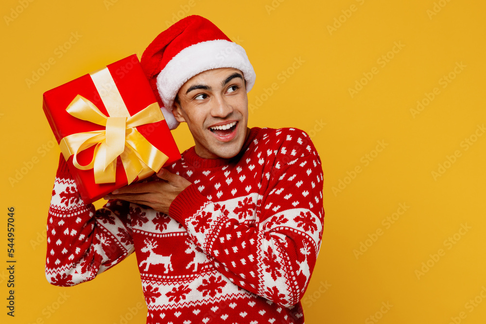 Merry fun young man wear christmas sweater Santa hat posing hold red present box with gift ribbon bow look aside on workspace isolated on plain yellow background. Happy New Year 2023 holiday concept. Merry fun young man wear christmas sweater Santa hat posing hold red present box with gift ribbon bow look aside on workspace isolated on plain yellow background. Happy New Year 2023 holiday concept.
